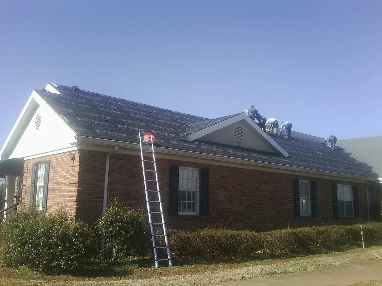 Crew tearing shingles off a brick ranch roof with a ladder set — Metal Roof Repair work by our crew in Horse Branch
