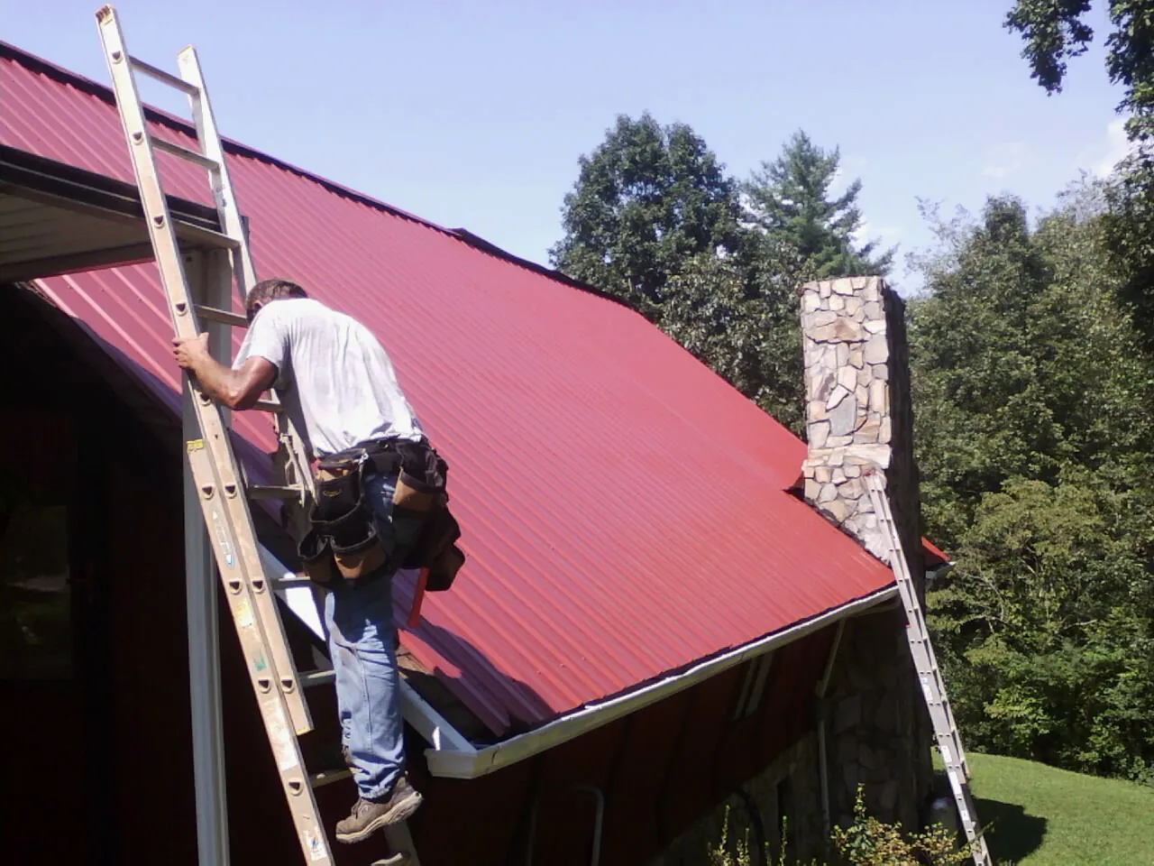 Roofer on a ladder beside a red metal roof and stone chimney — Soffit & Fascia Repair by Roofing Brothers in Horse Branch